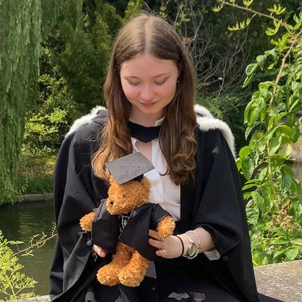 Louisa Yapp in a black graduation robe holding an orange Trinity Hall teddybear with greenery in the background.
