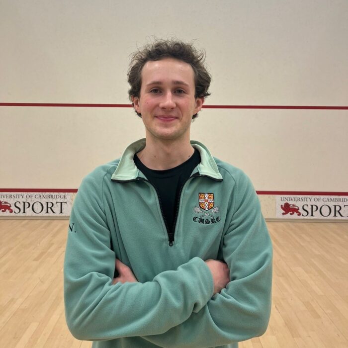Ollie Norton wearing a blue Cambridge University squash club sweatshirt standing in a squash court.